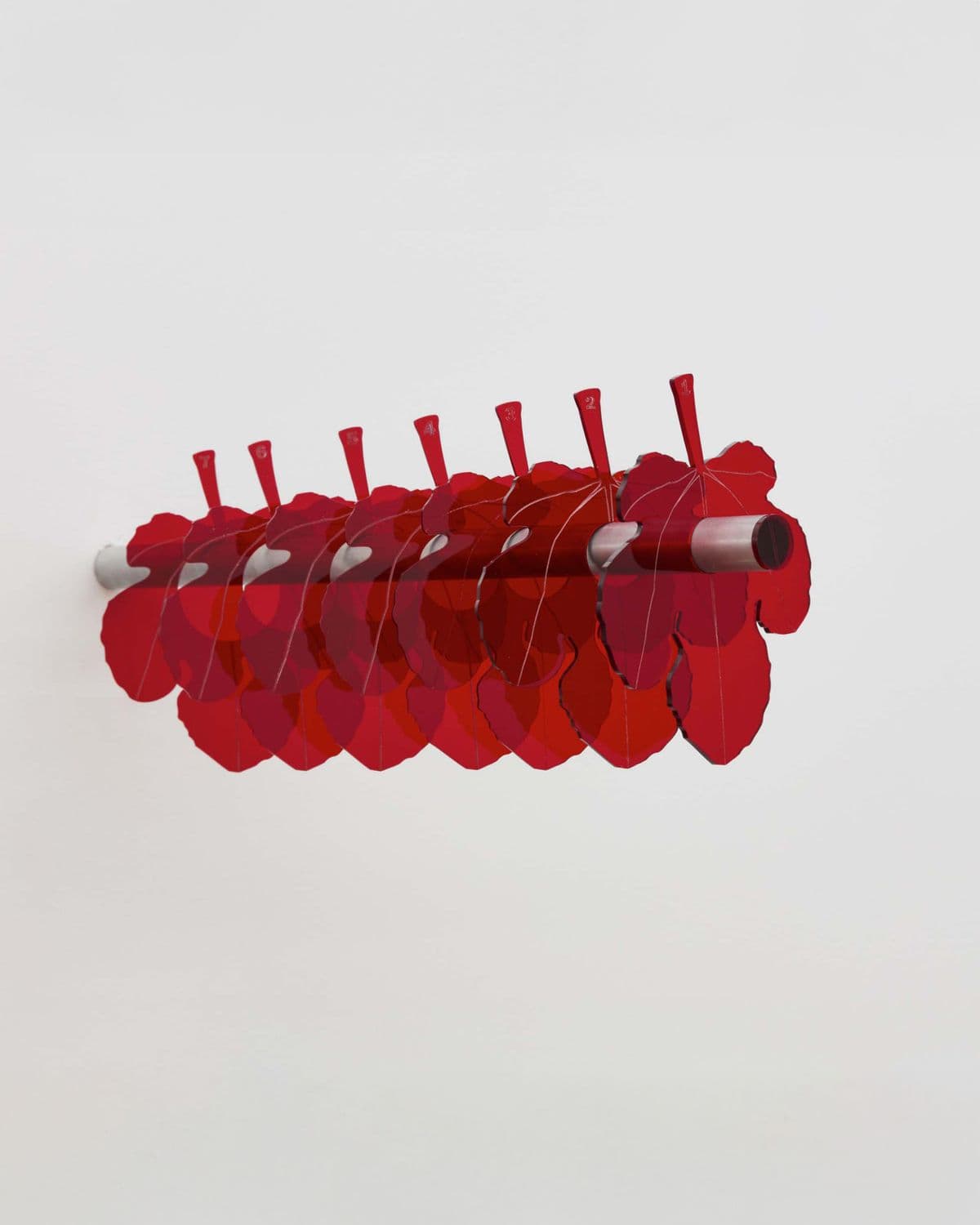 7x360°, wall-mounted sculpture by Grigorios Ilaridis featuring seven transparent red acrylic fig leaves suspended from a horizontal metal rod.