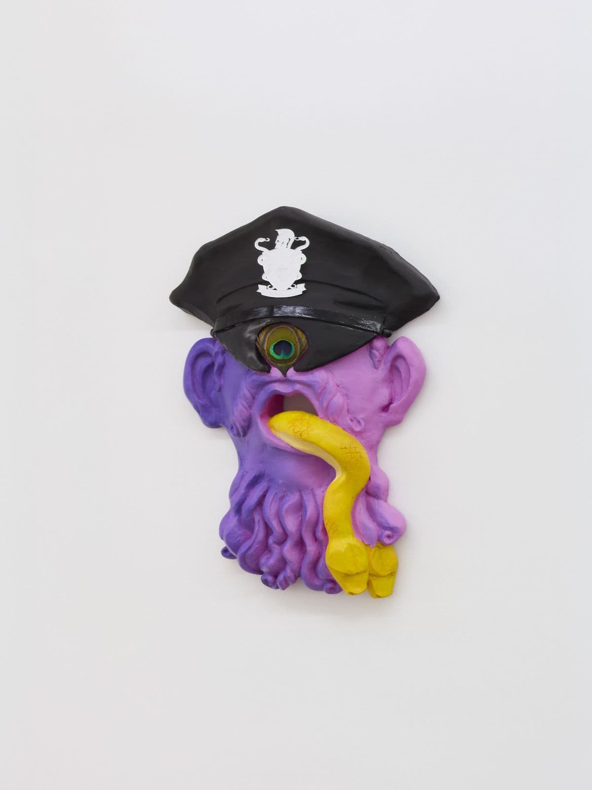 Oral Tradition I, wall-mounted Cyclops mask sculpture by Grigorios Ilaridis with a peacock feather eye, police cap and serpent emerging from the mouth. 
