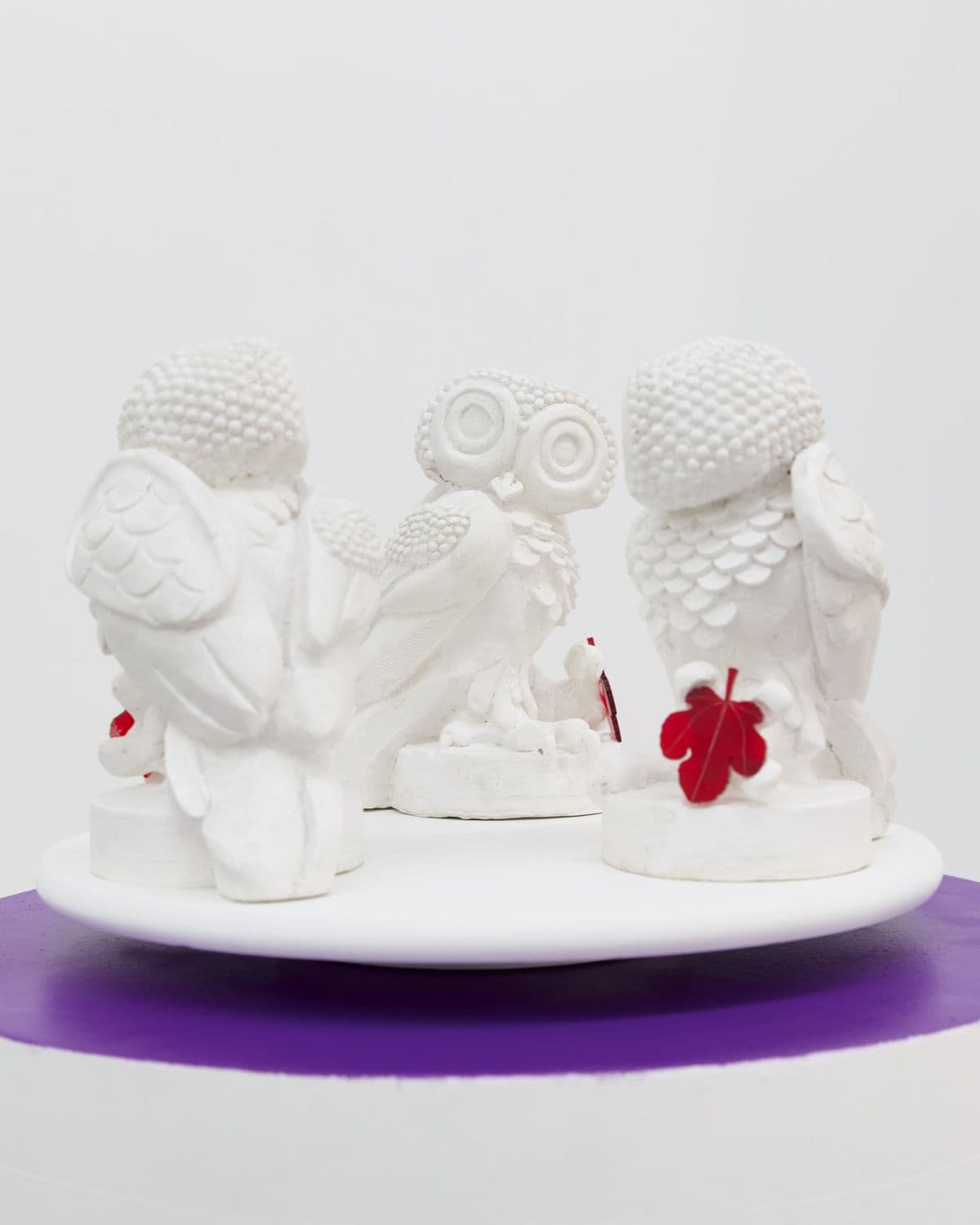 Wisdom in Orbit, sculpture by Grigorios Ilaridis featuring three white owls arranged in a circle, each holding a transparent red acrylic fig leaf.